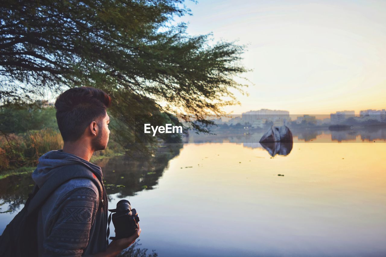 Man holding camera while standing by lake against sky during sunset