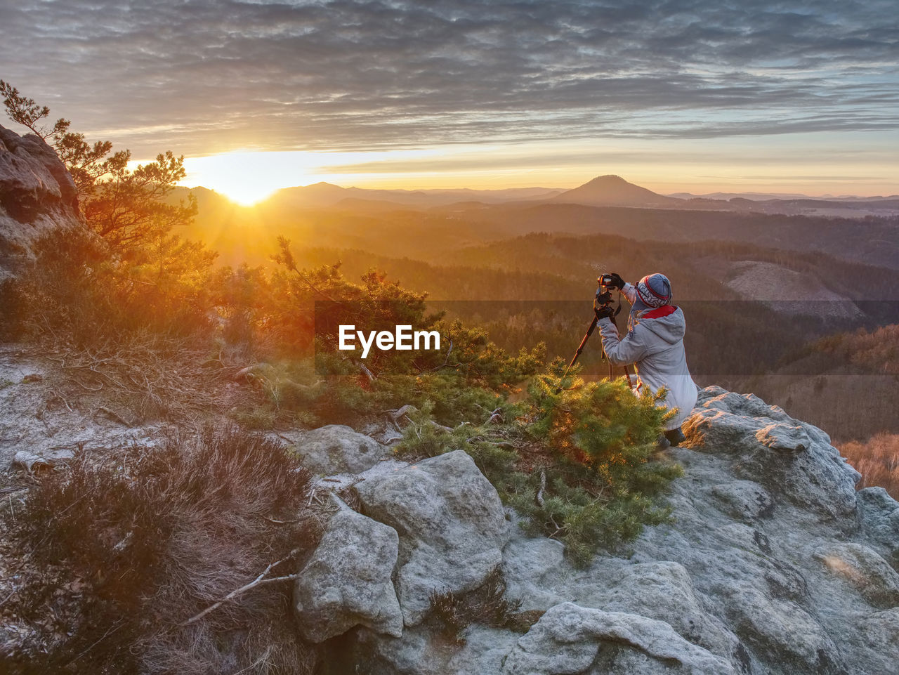 Woman hiker and photographer set tripod with camera on exposed rock above deep misty valley
