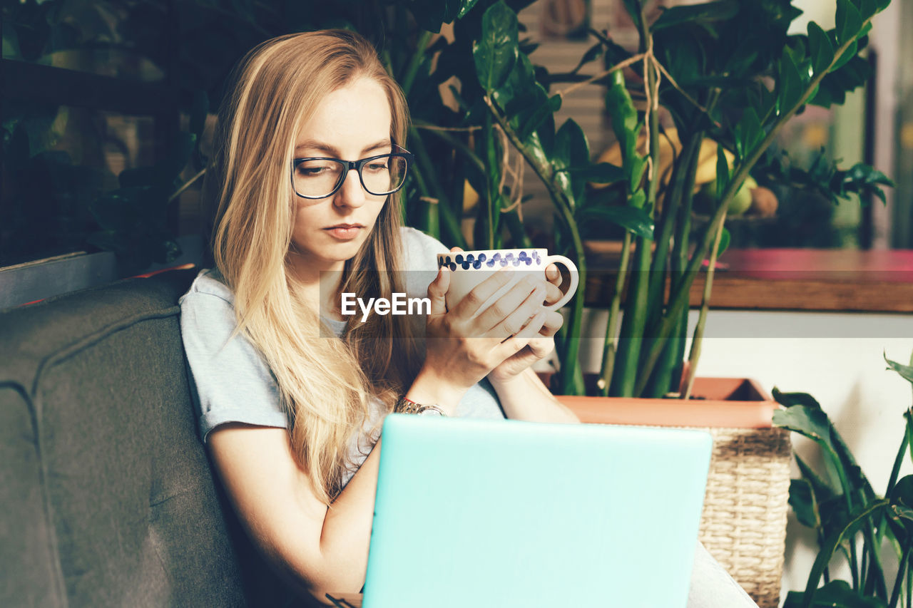 A student girl in glasses is holding a coffee mug and surfing the internet in a laptop.