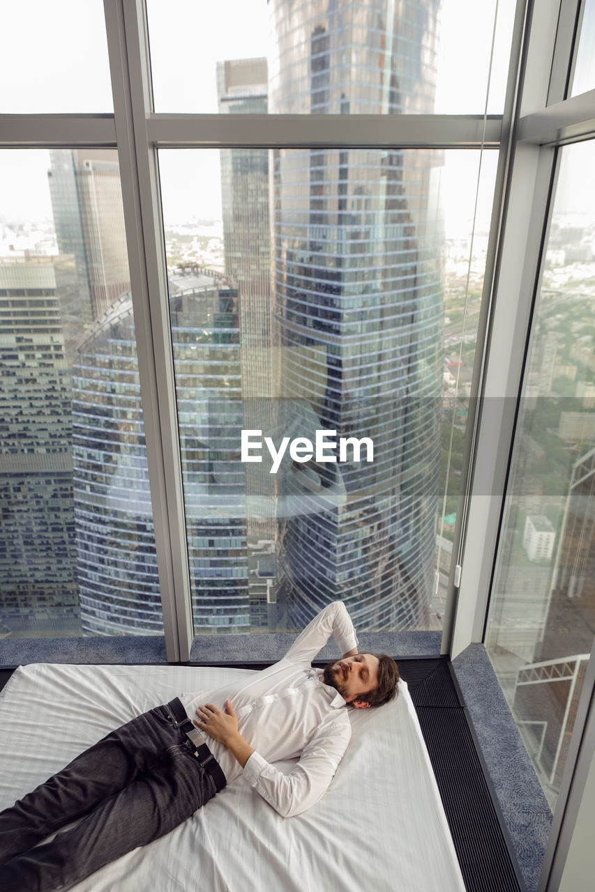 window, adult, one person, indoors, interior design, architecture, men, glass, city, lying down, day, relaxation, looking through window, built structure, lifestyles, building, window covering, young adult, business, office building exterior, cityscape, transparent, office, businessman, women, skyscraper, communication, looking, wireless technology, mature adult, furniture, full length