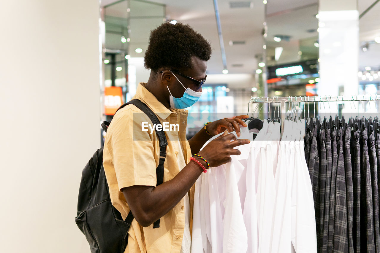 Side view of african american male in medical mask standing in clothing store and choosing t shirt during coronavirus epidemic