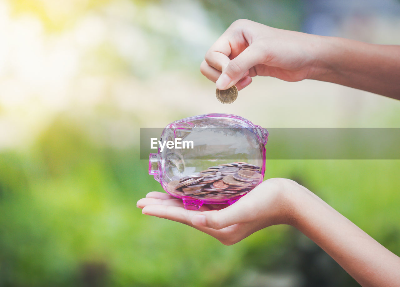 Close-up of person holding coin over transparent piggy bank 