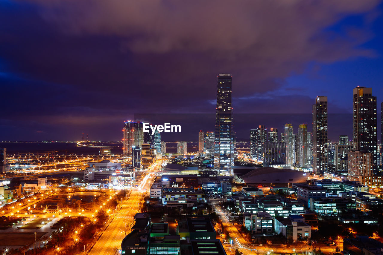 Light trails on street by illuminated cityscape against cloudy sky at dusk