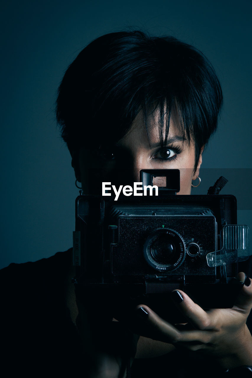 Concentrated youthful dark haired female model looking at camera and photographing with retro camera in studio on blue background