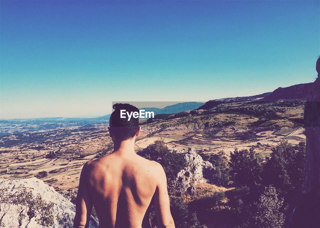 REAR VIEW OF SHIRTLESS MAN STANDING ON MOUNTAIN AGAINST CLEAR SKY