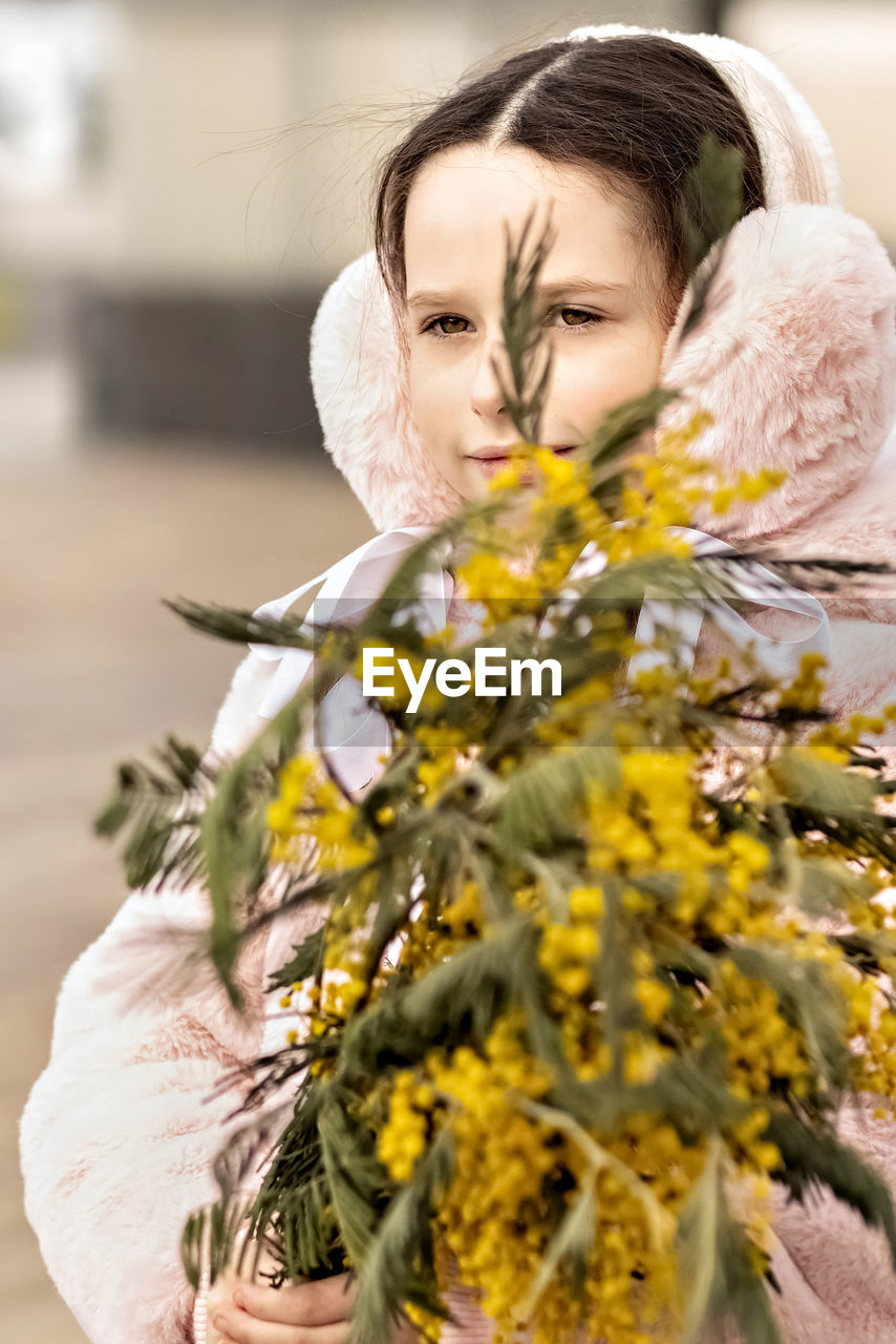 Portrait of a toddler girl in a warm pink coat and fur headphones with a bouquet of mimosa 