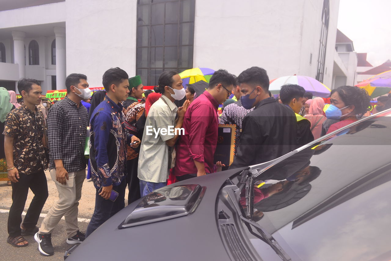 group of people, men, car, architecture, transportation, mode of transportation, adult, vehicle, motor vehicle, crowd, women, city, large group of people, building exterior, day, clothing, built structure, togetherness, land vehicle, standing, outdoors, person, young adult