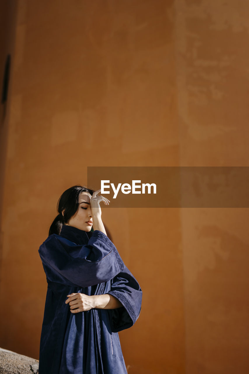 Young woman with eyes closed and head in hands in front of orange wall on sunny day