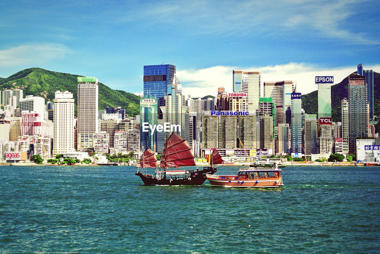 Boats sailing in sea by cityscape against sky