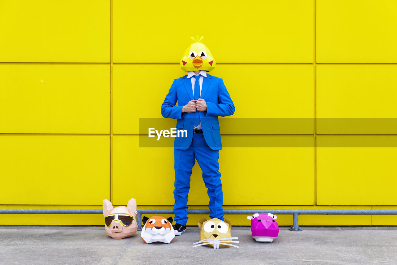 Man wearing vibrant blue suit and bird mask standing in front of yellow wall with various animal masks