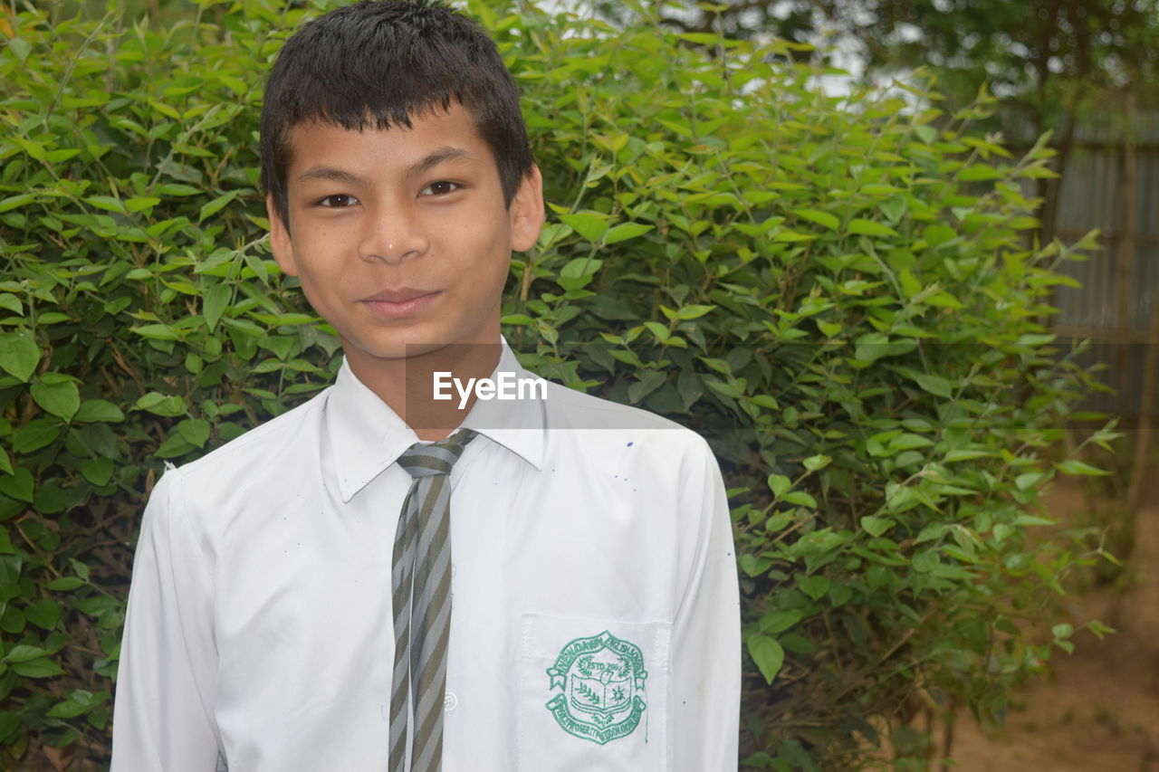Portrait of smiling young man wearing school uniform standing against plants