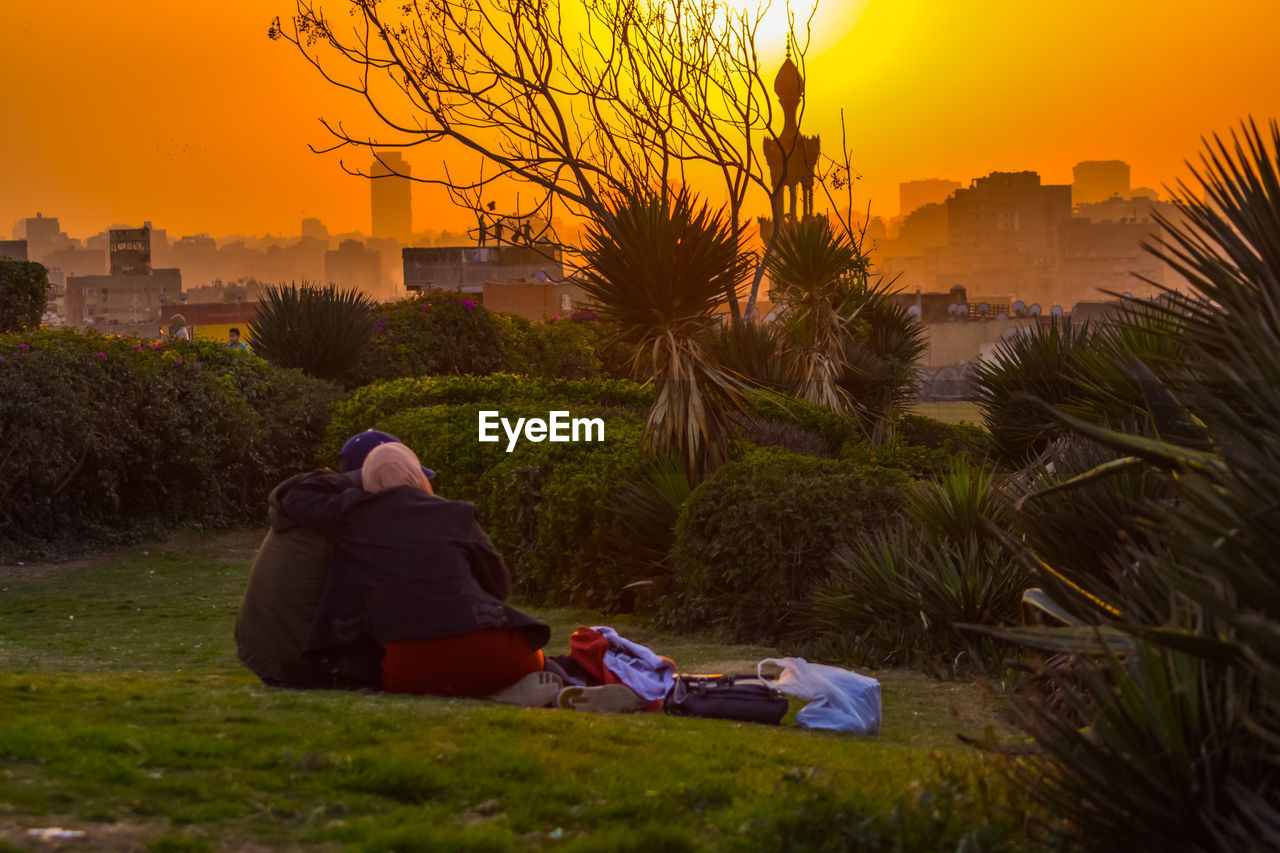 REAR VIEW OF PEOPLE SITTING AT PARK AGAINST SKY