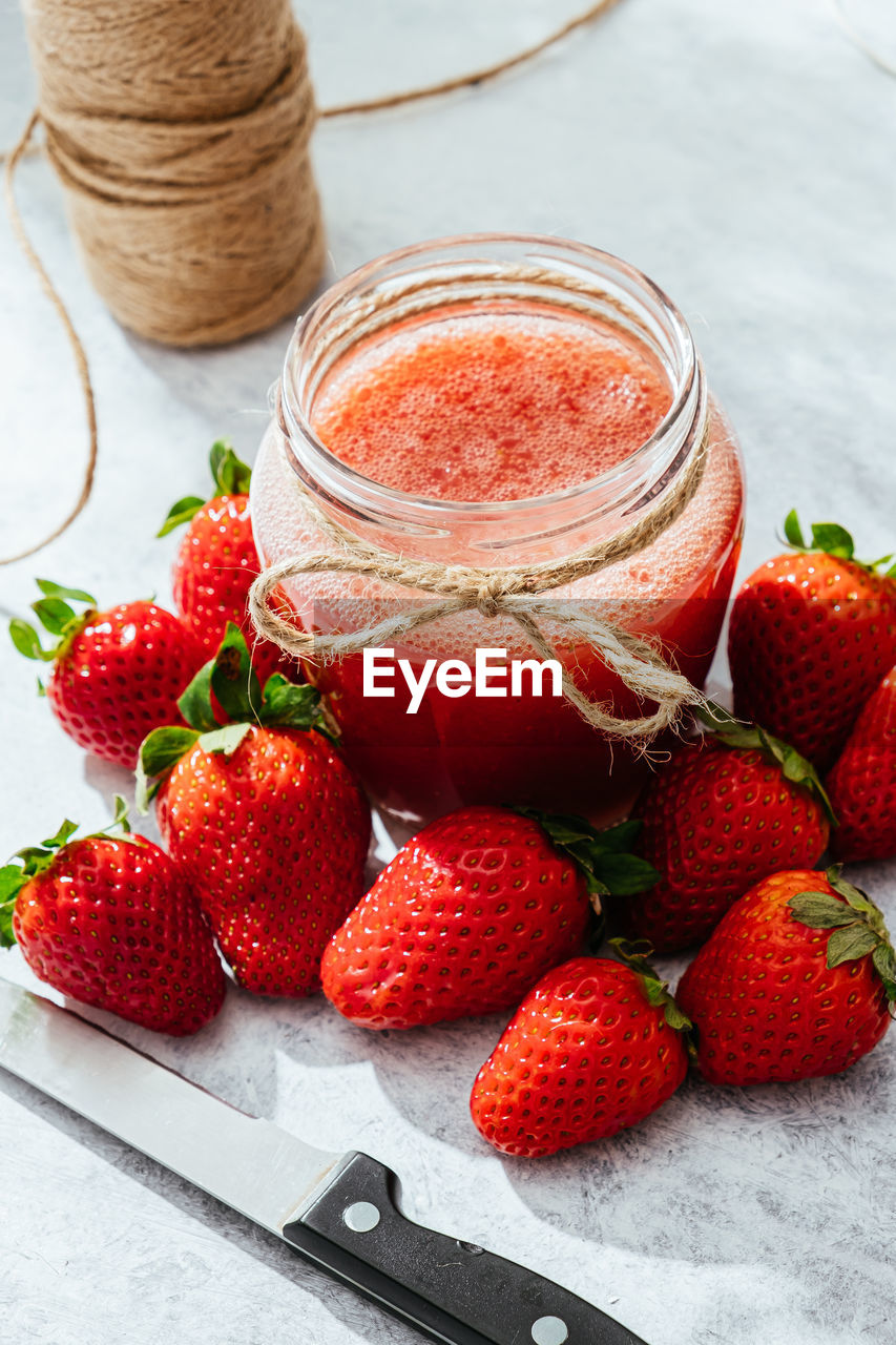 Composition with fresh homemade strawberry juice in glass jar wrapped with twine placed on marble surface with whole berries and knife