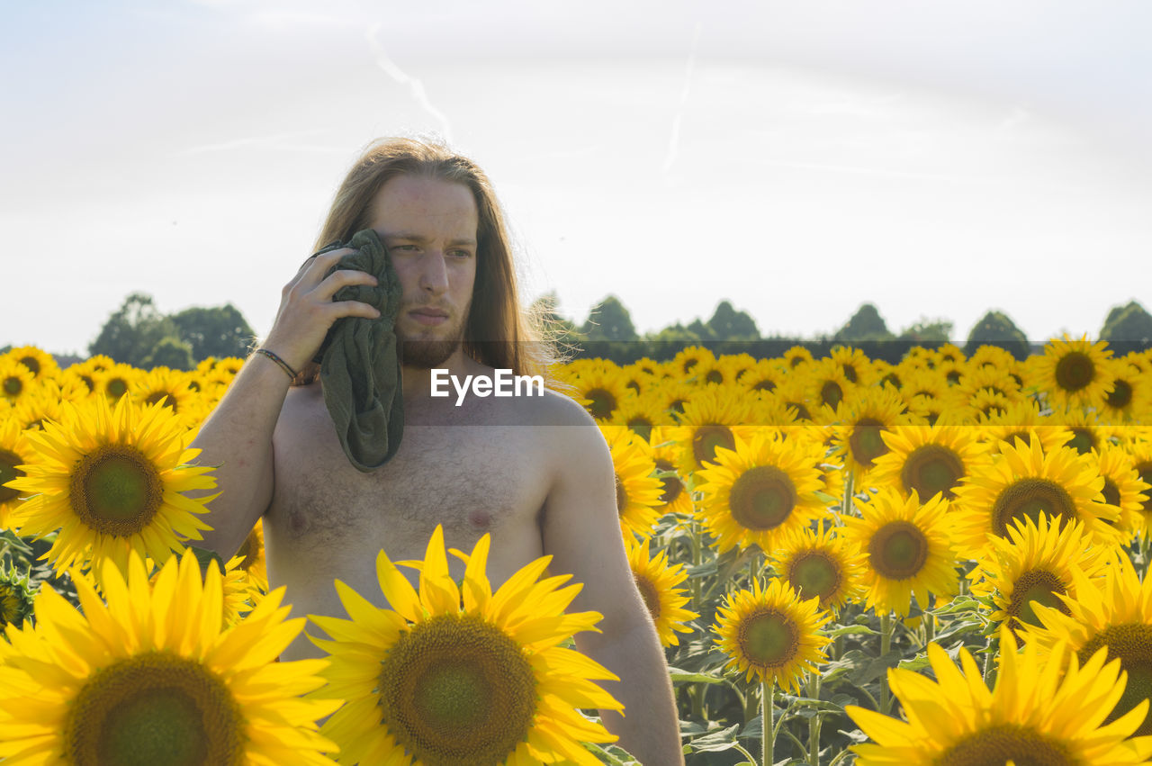 Thoughtful shirtless man wiping himself from towel while standing amidst sunflowers