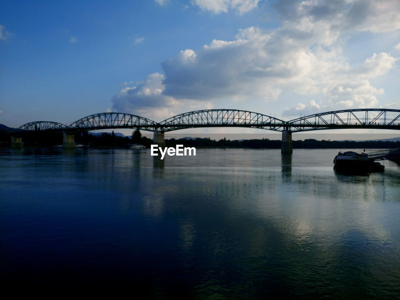 BRIDGE OVER CALM RIVER AGAINST CLOUDY SKY