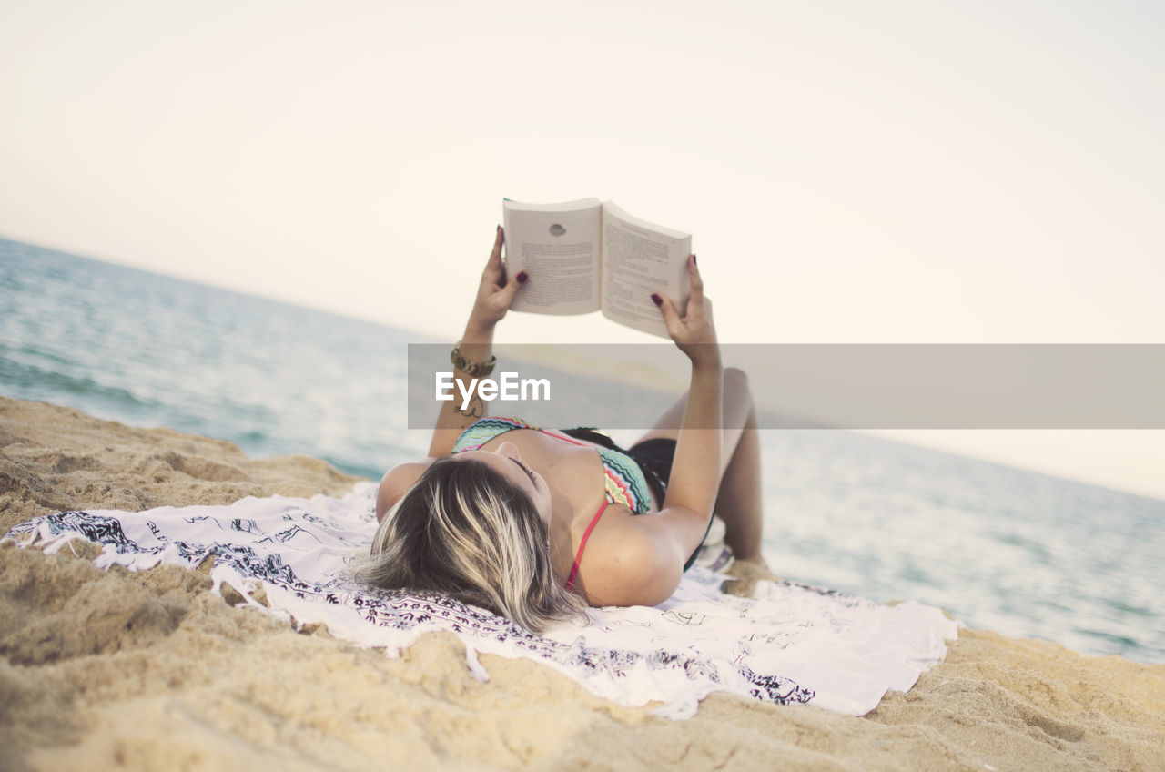 Woman reading book while relaxing at beach against sky