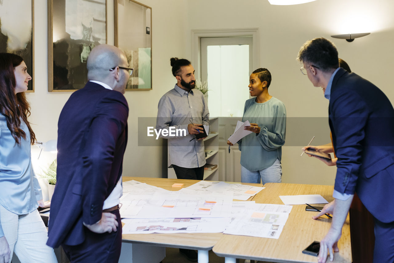 Confident female entrepreneur discussing with colleagues over blueprint at table in office