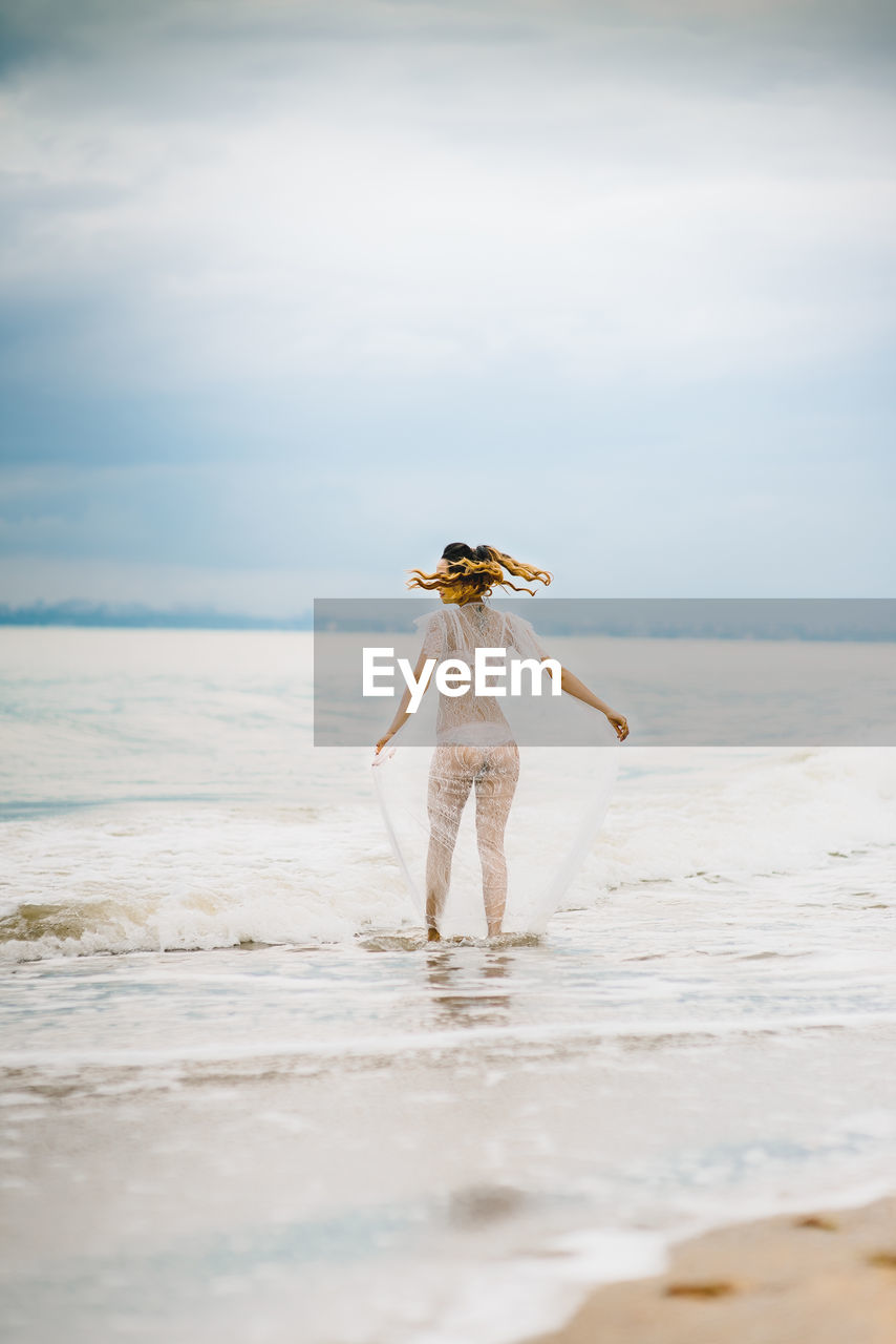 REAR VIEW OF WOMAN STANDING ON BEACH AGAINST SEA