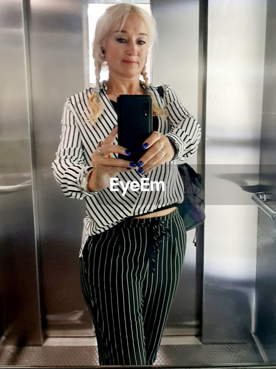 one person, striped, adult, women, wireless technology, technology, communication, smartphone, mobile phone, indoors, portable information device, holding, standing, business, female, blond hair, clothing, dress, business finance and industry, looking, front view, businesswoman, telephone, mature adult, using phone, fashion, person, lifestyles, casual clothing, convenience, digital tablet, portability, elevator