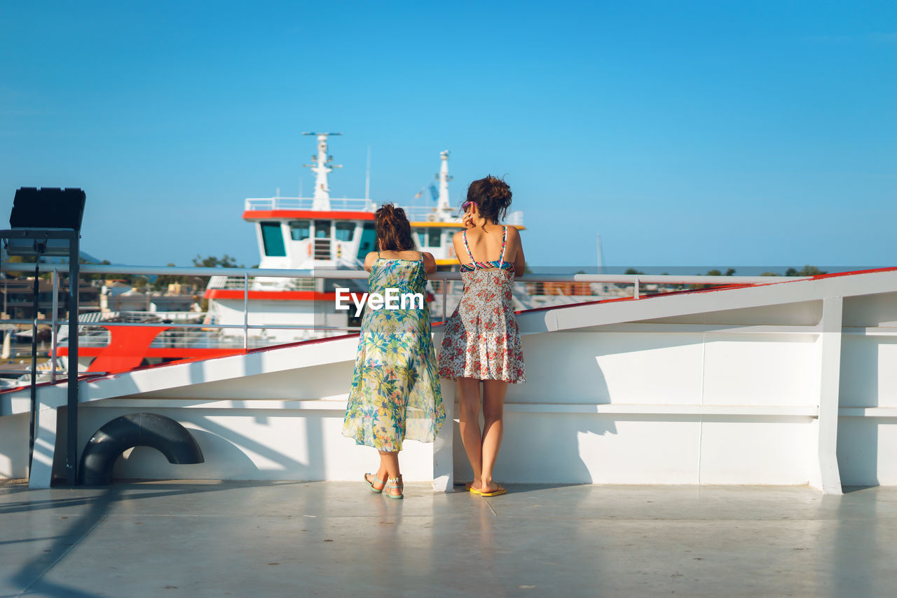 Rear view of women standing by railing on boat against blue sky