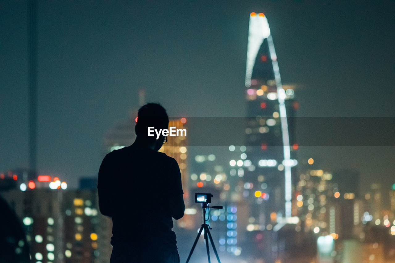 Silhouette man standing by digital camera on tripod against illuminated cityscape at night