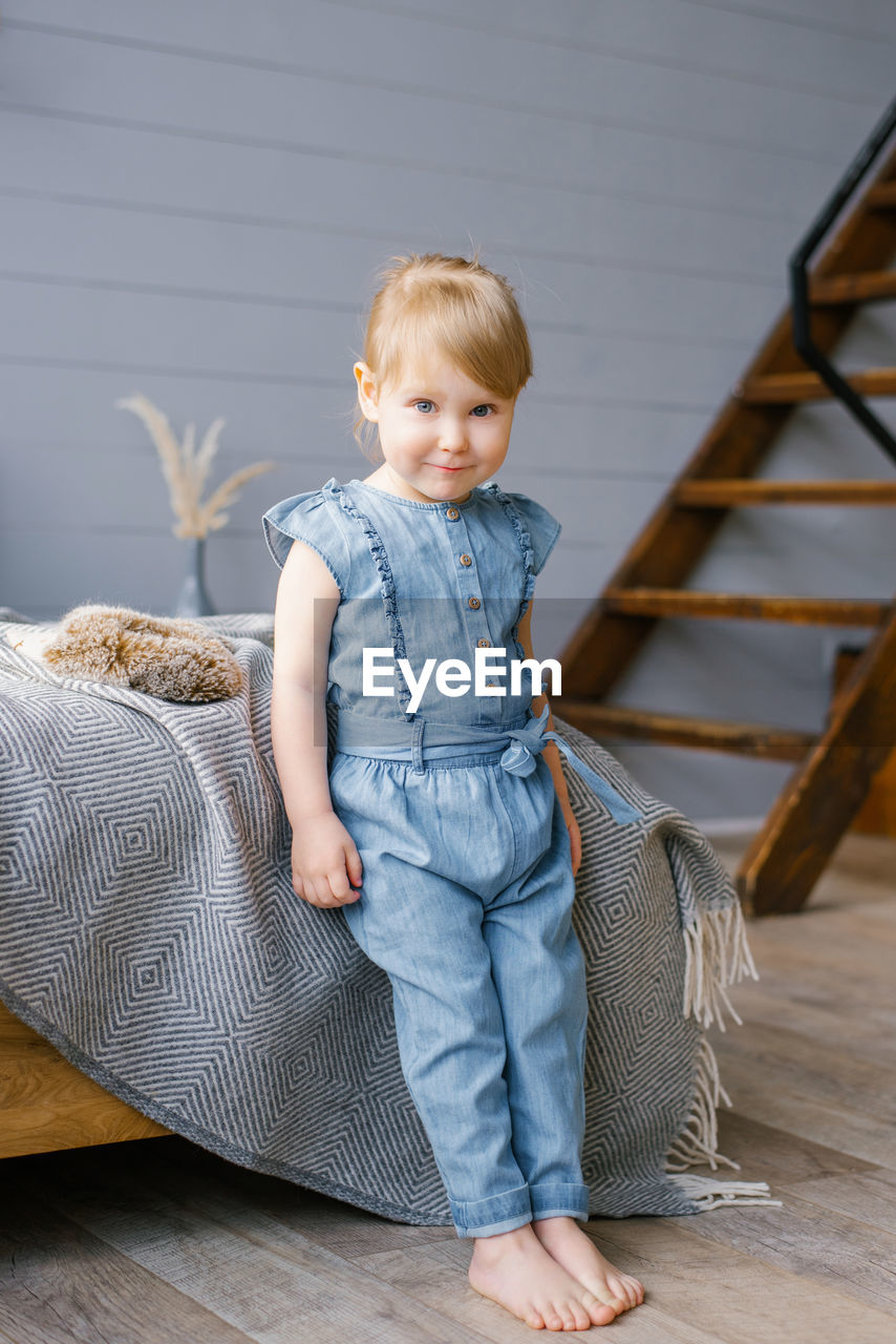 A cute two-year-old girl in a denim jumpsuit with bare feet stands by the bed at home