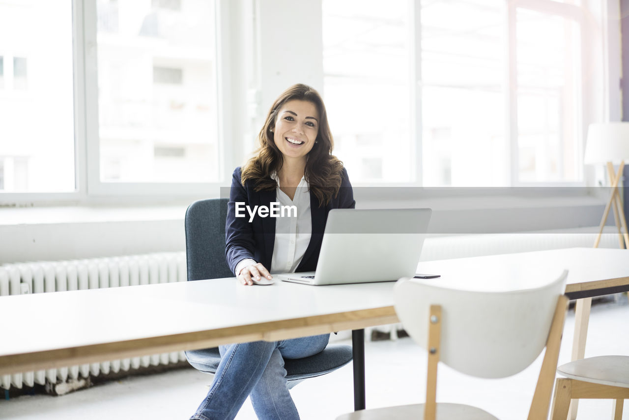 Portrait of happy businesswoman sitting at desk in the office working on laptop