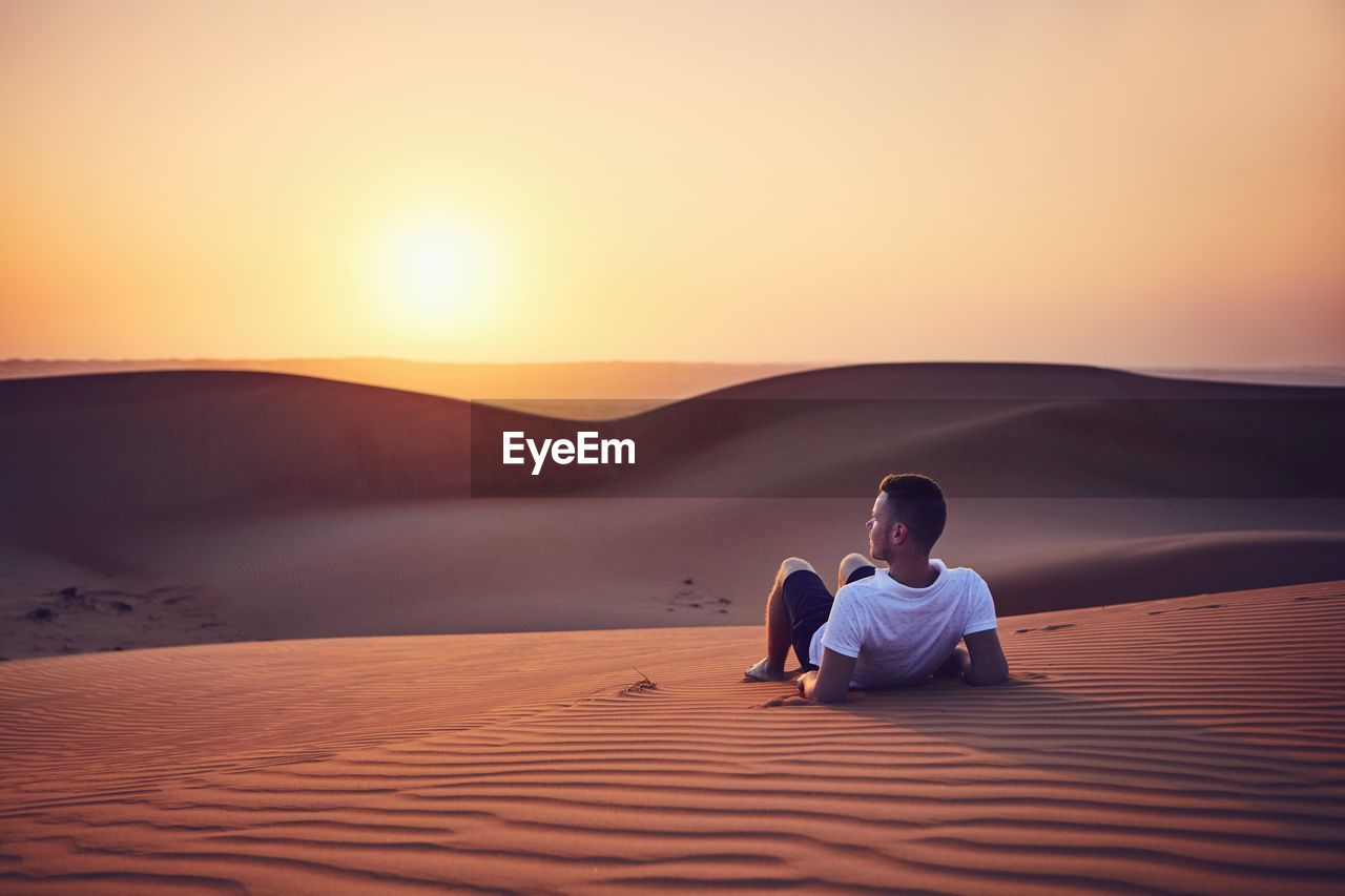 Man leaning on sand dune in desert against sky during sunset