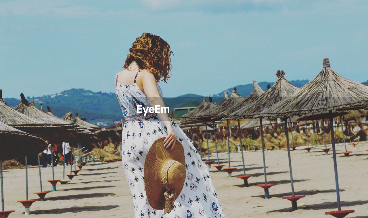 Rear view of woman holding hat while standing at beach against sky