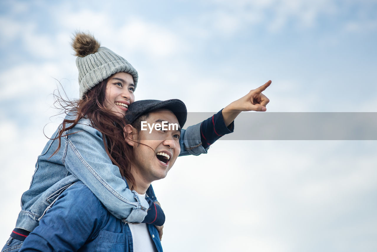 PORTRAIT OF HAPPY YOUNG WOMAN WITH ARMS RAISED AGAINST SKY