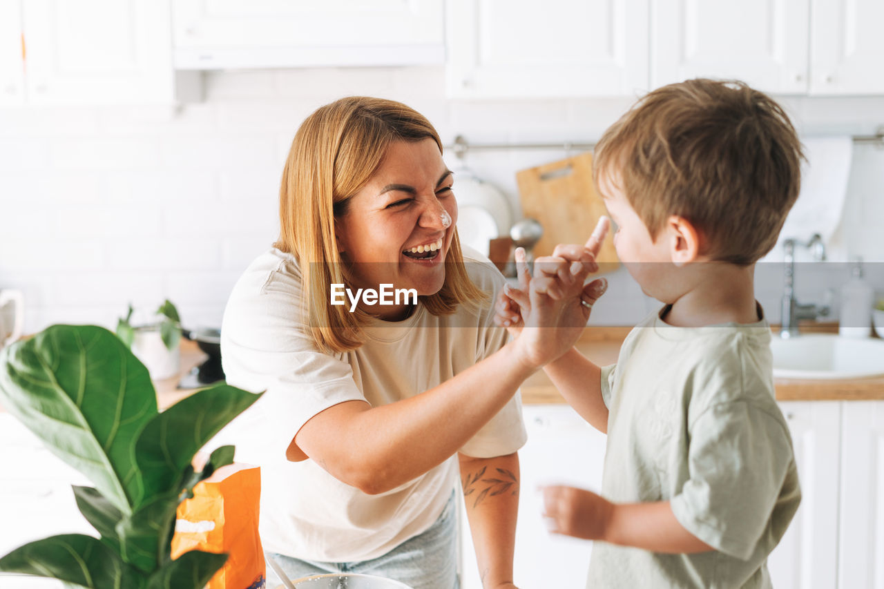 Young woman mother and toddler boy son have fun while cooking with flour at table in bright kitchen 