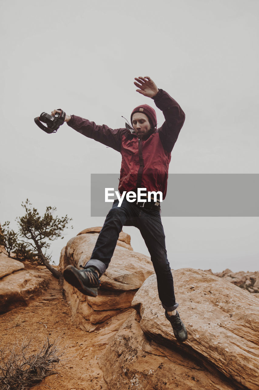 Low angle view of man holding camera while jumping on rocks against sky