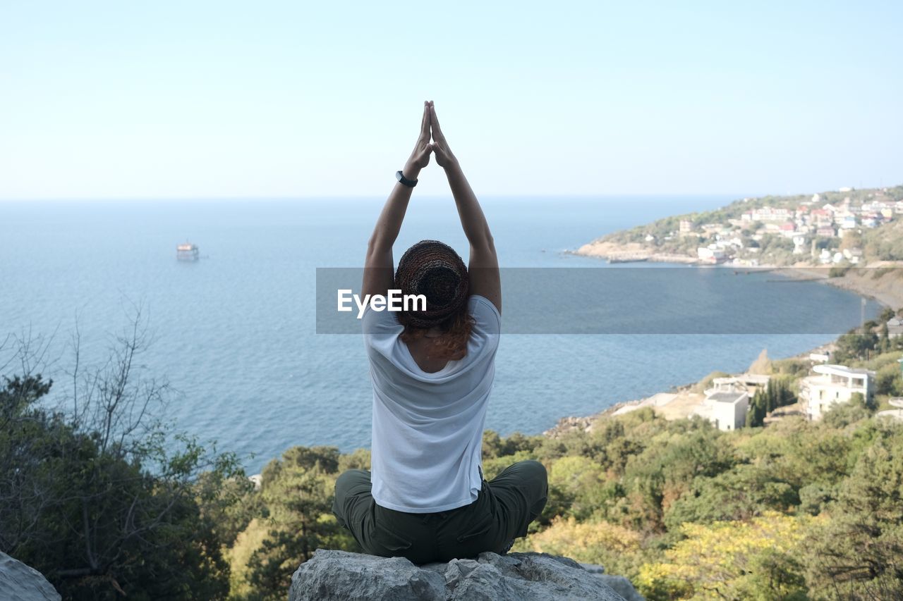 Rear view of woman meditating with arms raised on rock against sea