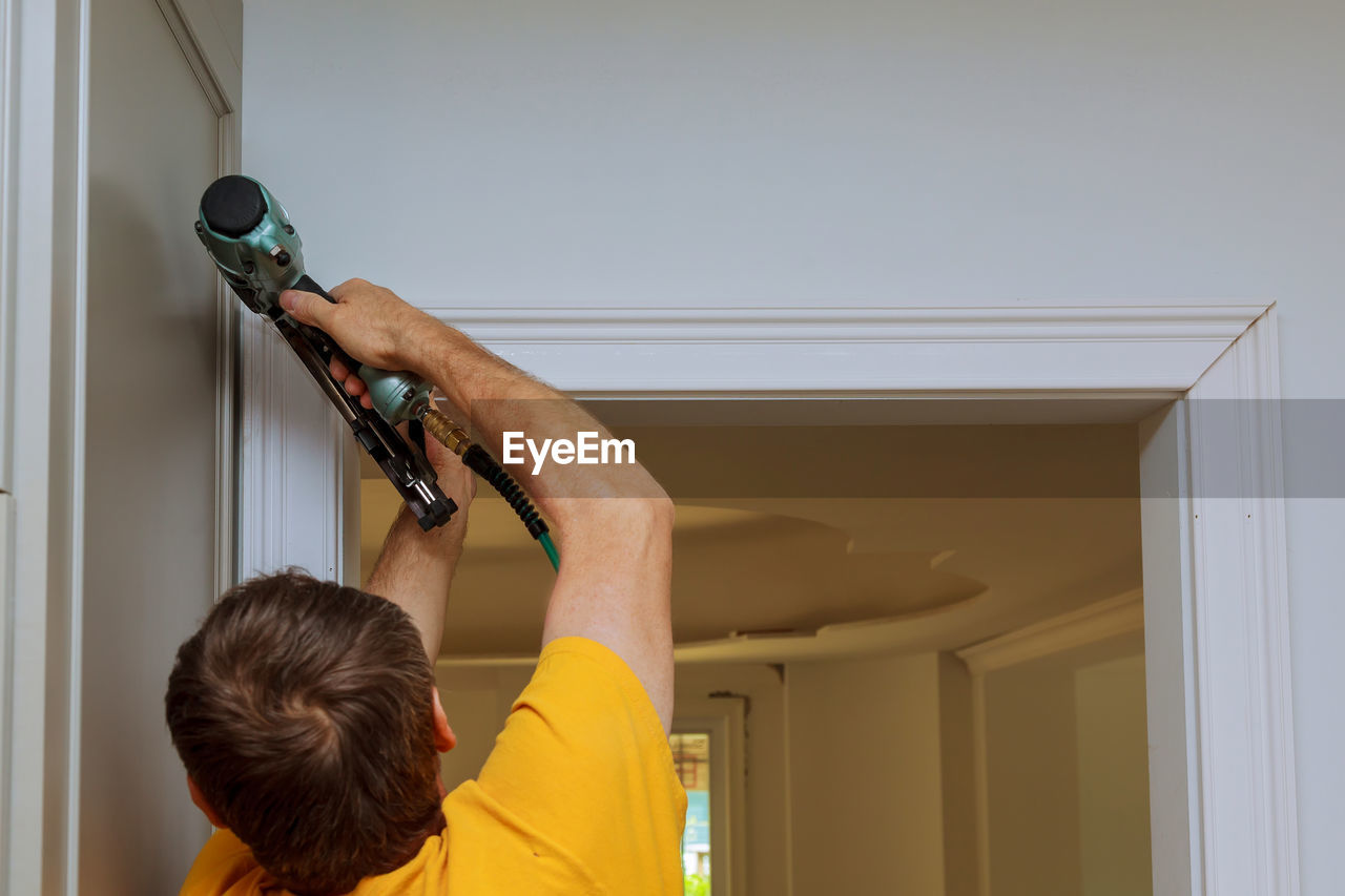 Rear view of man using nail gun on wall at home