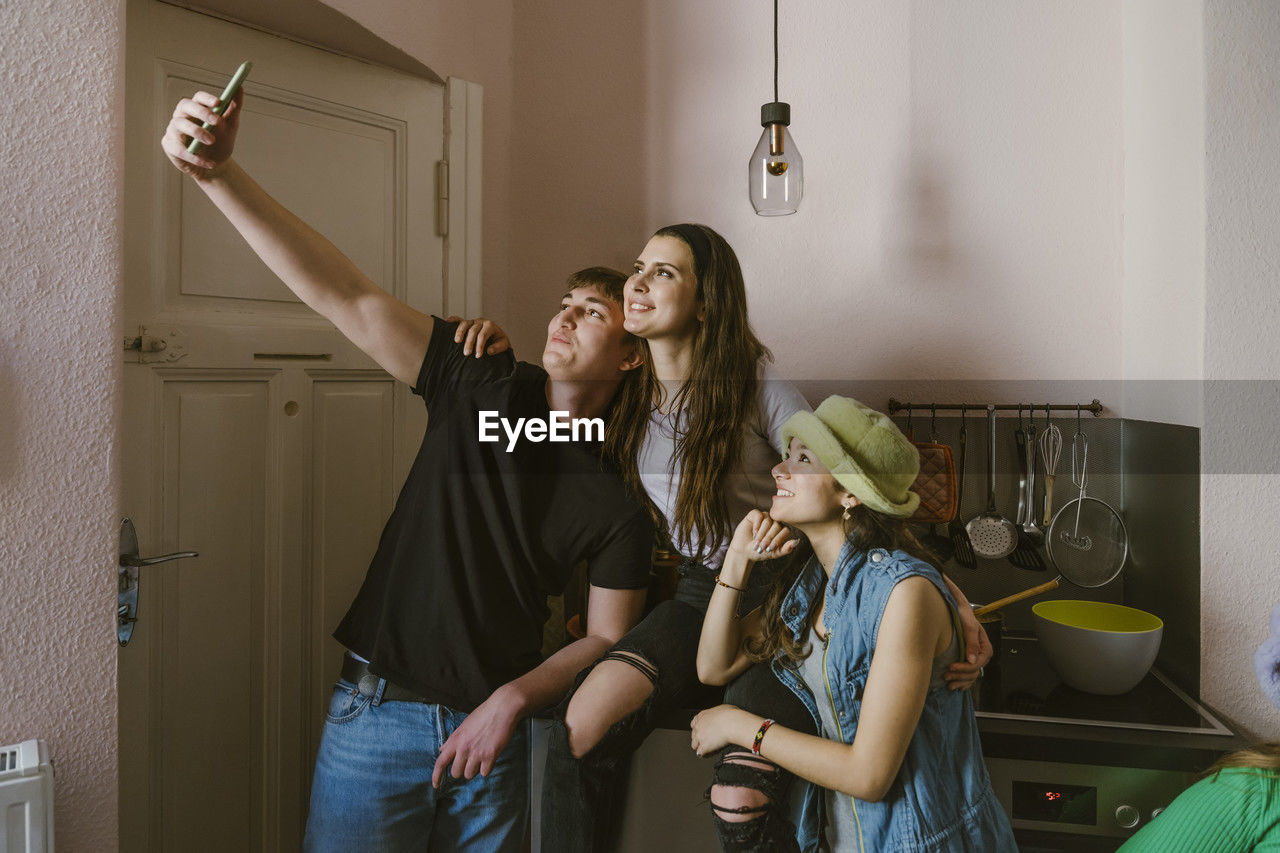 Young man taking selfie with smiling female friends in kitchen at home