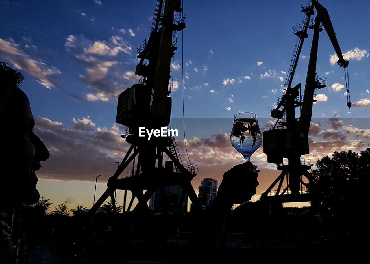 Man holding drink glass by cranes during sunset