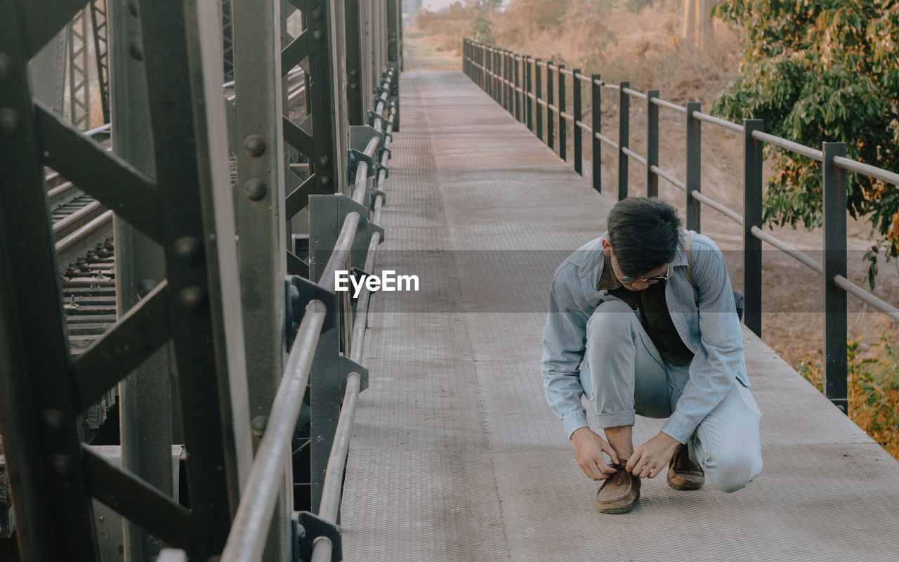 Full length of young man tying shoelace on bridge