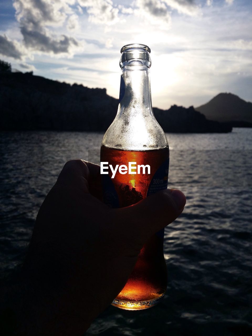 Cropped image of person holding drink in bottle by sea against sky