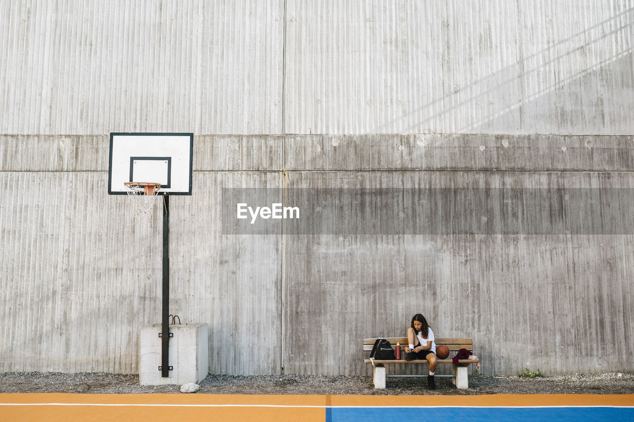 Girl tying shoelace while sitting on bench at basketball court