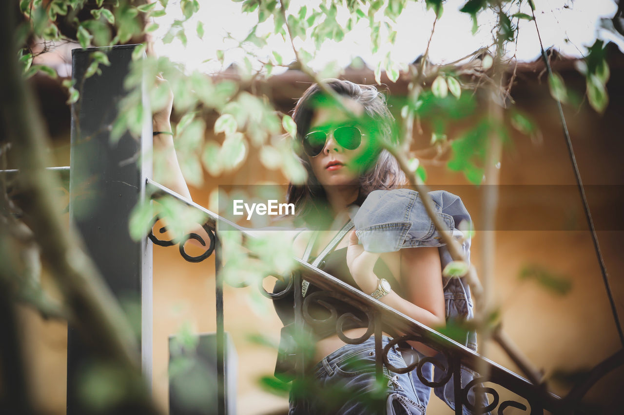 Portrait of young woman holding wearing sunglasses standing against railing