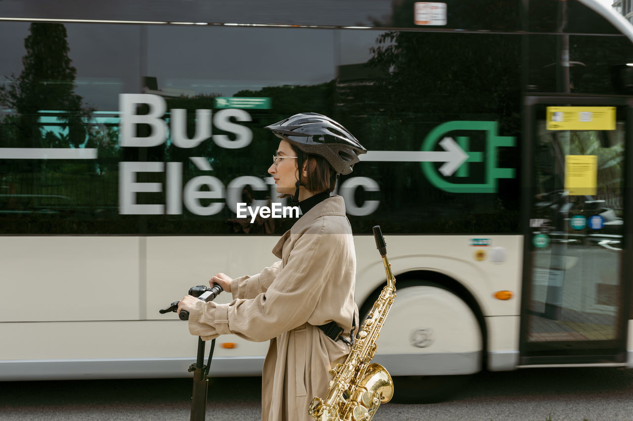 Woman carrying saxophone riding electric push scooter at city