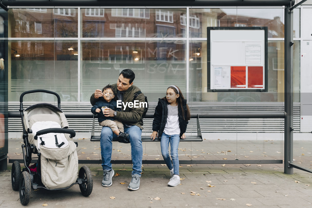 Father feeding baby food to son while sitting with daughter at bus stop in city