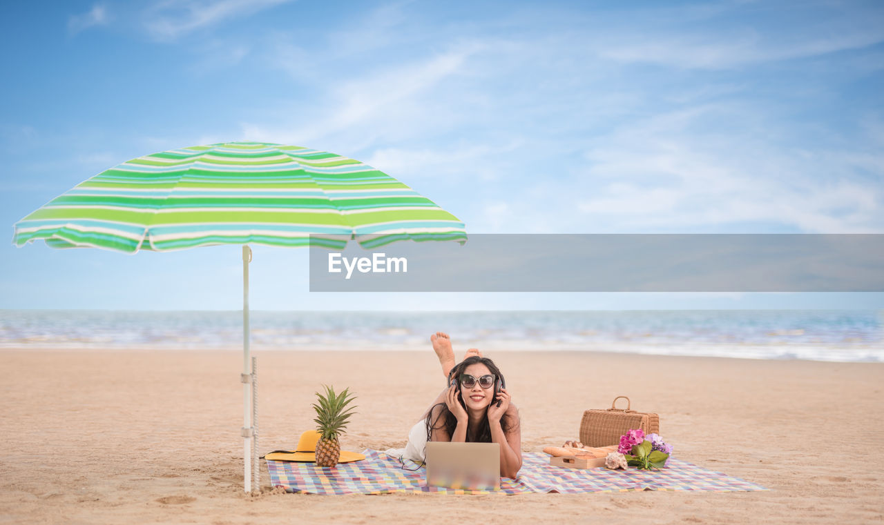 Woman with laptop enjoying music by parasol on beach against sky