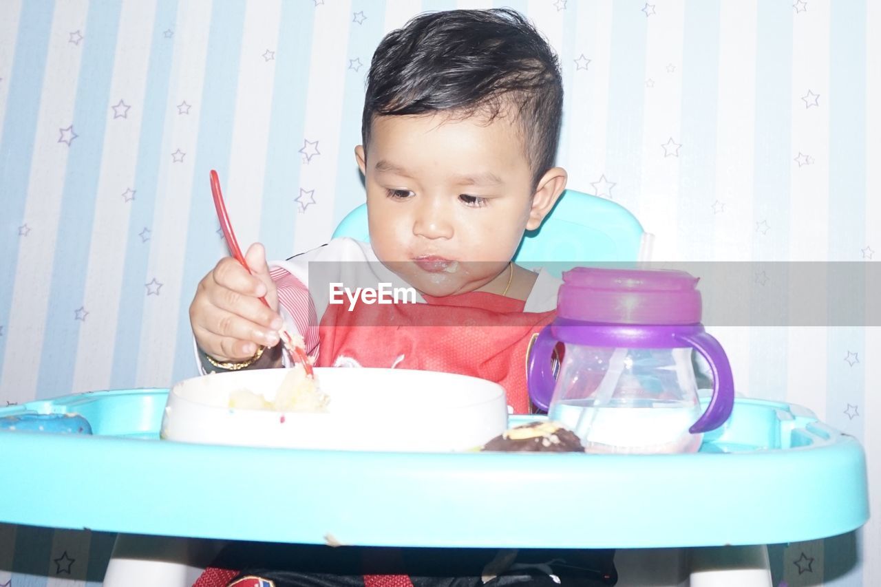 Close-up of cute baby boy eating food while sitting on high chair against wall 