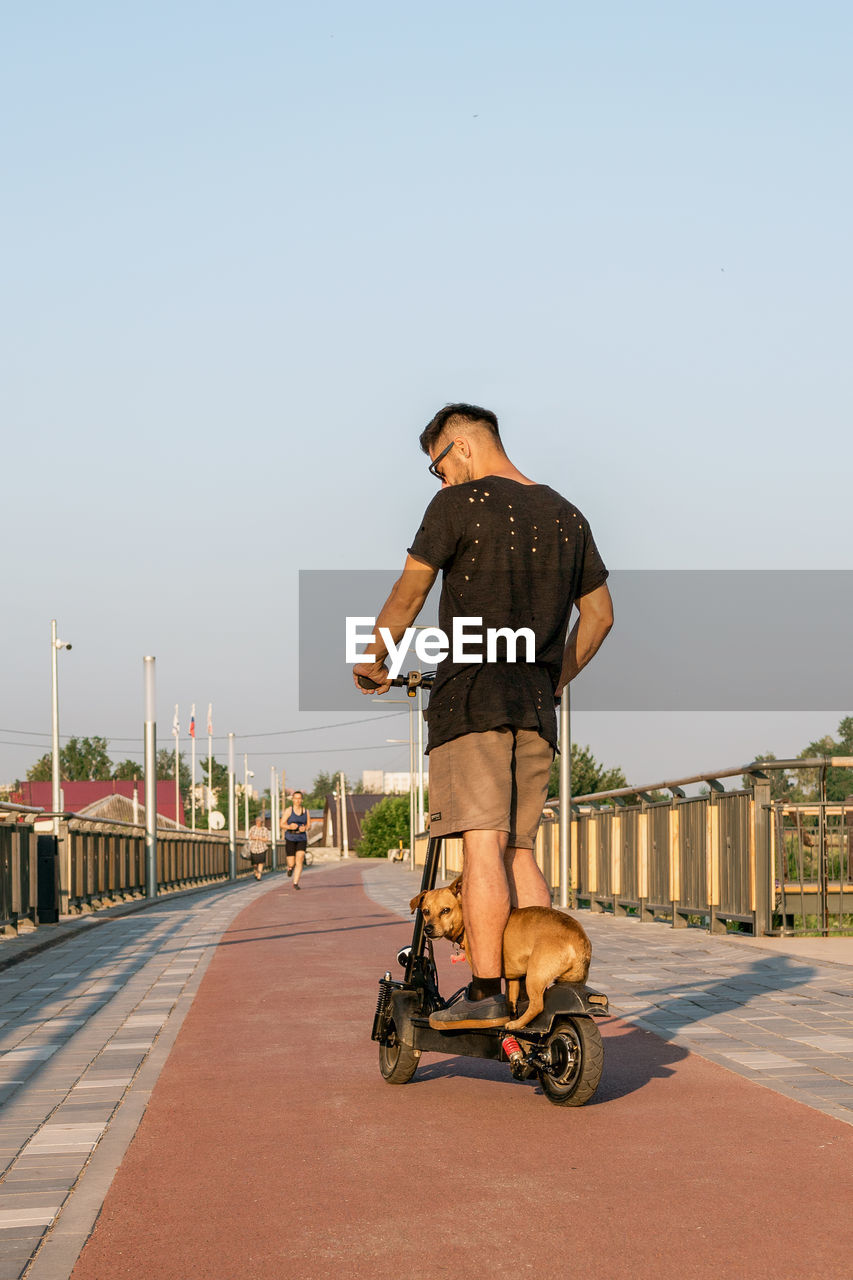 Man is riding on electric scooter with his small dog. walking with dog on empty street of city. 