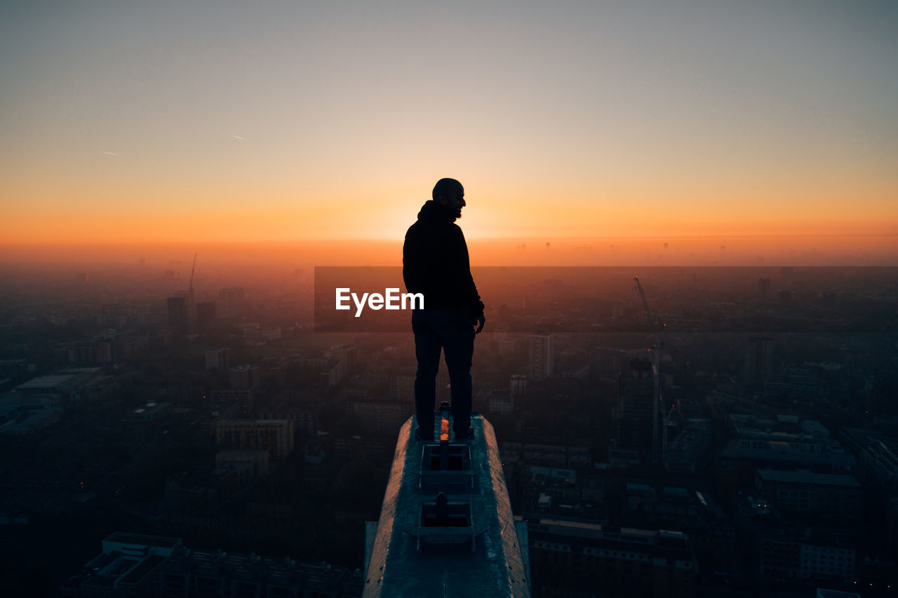 Rear view of silhouette man looking at cityscape against sky during sunset