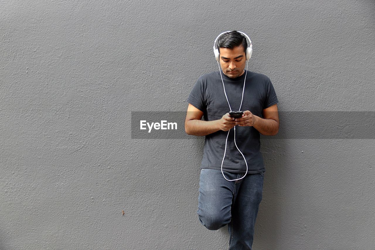 Young man listening music while using mobile phone against wall