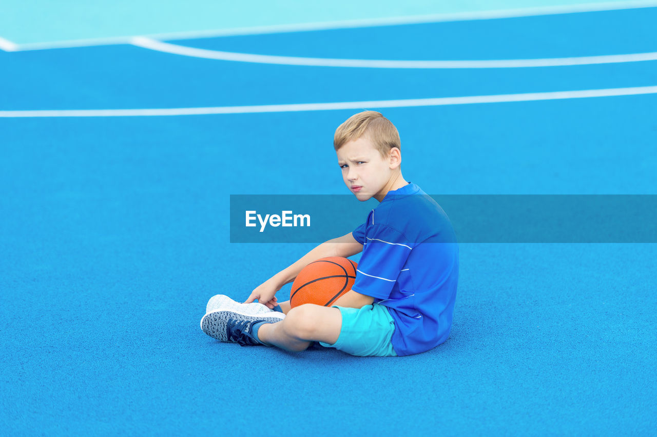 Portrait of boy sitting with basketball at court