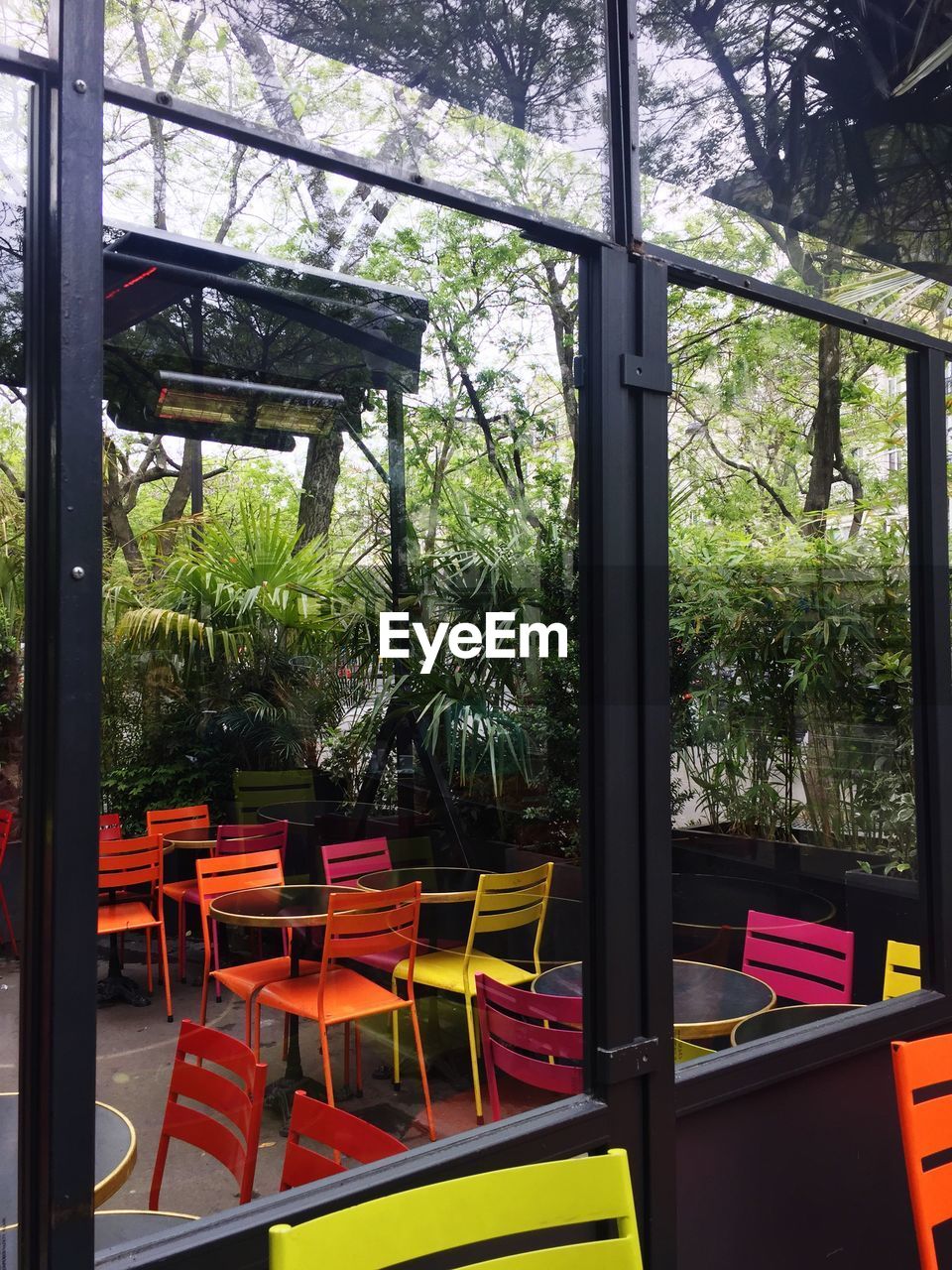 EMPTY CHAIRS AND TABLE AGAINST TREES SEEN THROUGH OPEN GLASS