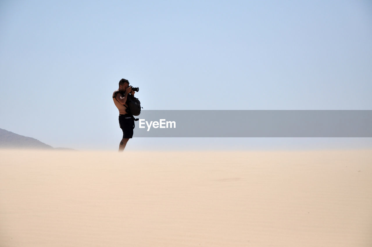 Young man photographing while standing in desert against clear sky